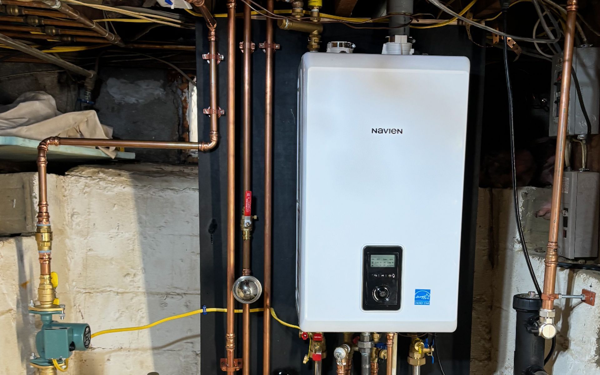 A white Navien tankless water heater mounted on a black panel in a basement, connected by various copper plumbing pipes.