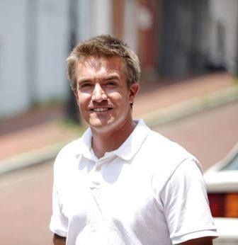 A smiling person with light brown hair wearing a white polo shirt outdoors on a paved street.