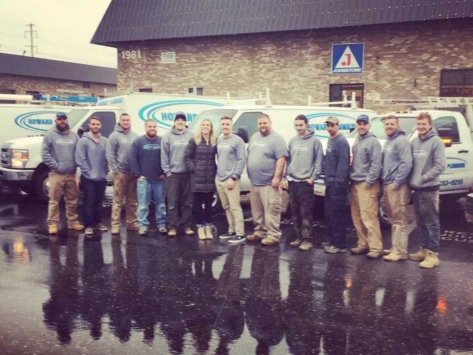 A team of workers in grey hoodies and tan pants stand in a row before a fleet of white service vans in a parking lot.