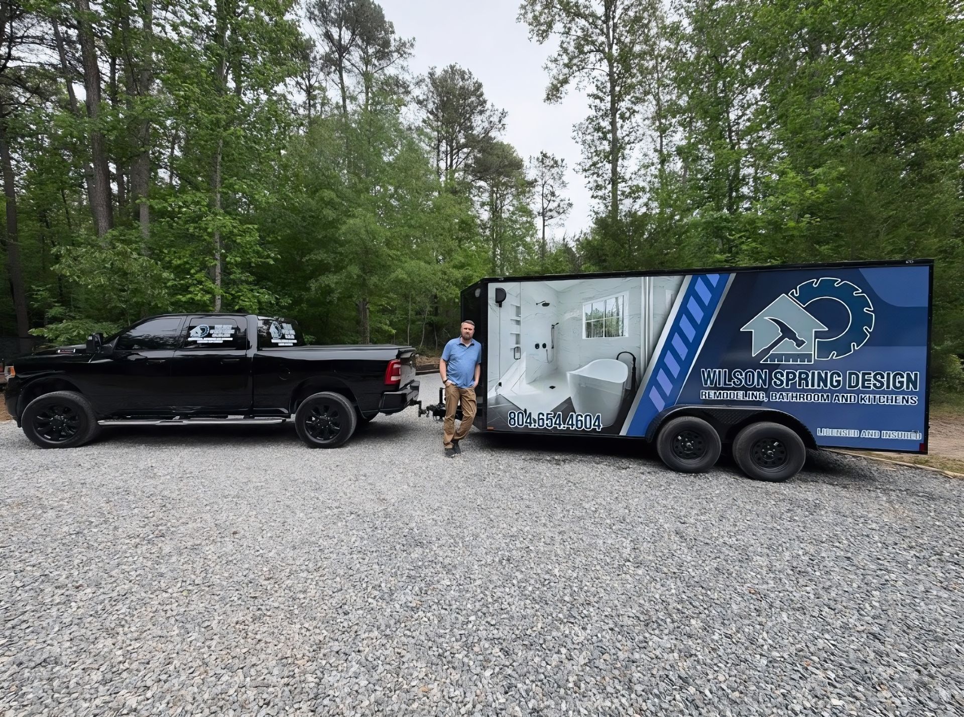 Black pickup truck hitched to a blue enclosed trailer with a person standing beside it on a gravel road in the woods