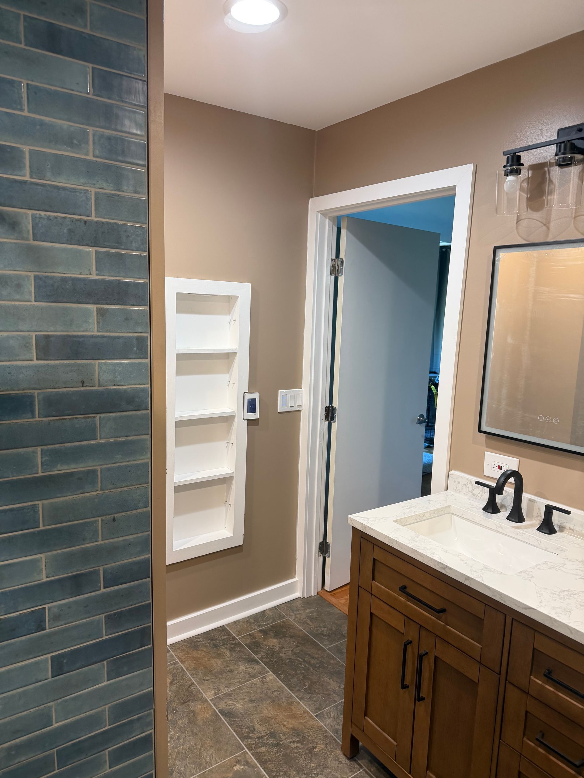 Bathroom interior with dark tile shower wall, beige walls, and a wooden vanity with mirror.