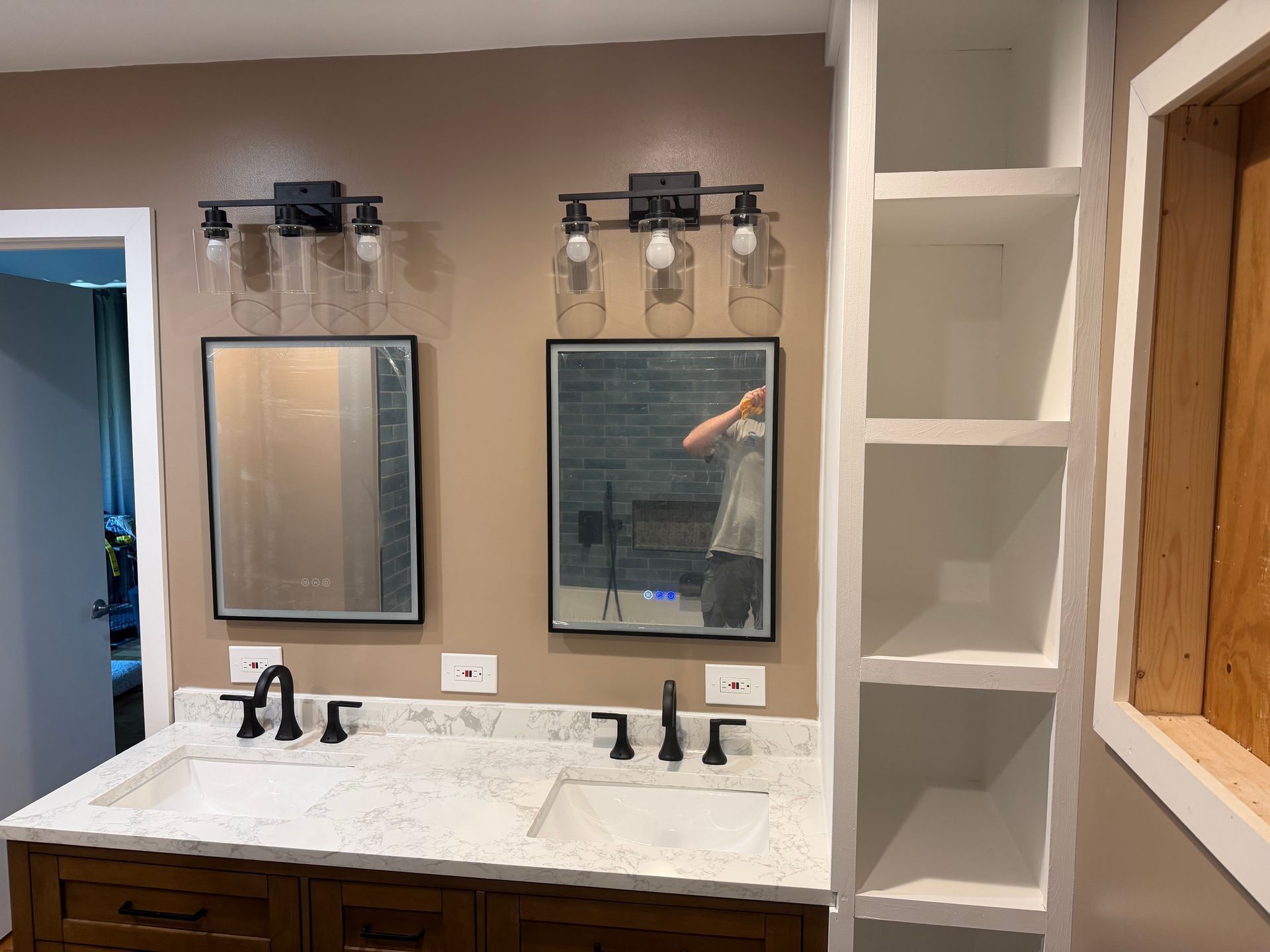 Bathroom vanity with double mirrors, black faucets, marble countertop, and built-in shelves beside a window.