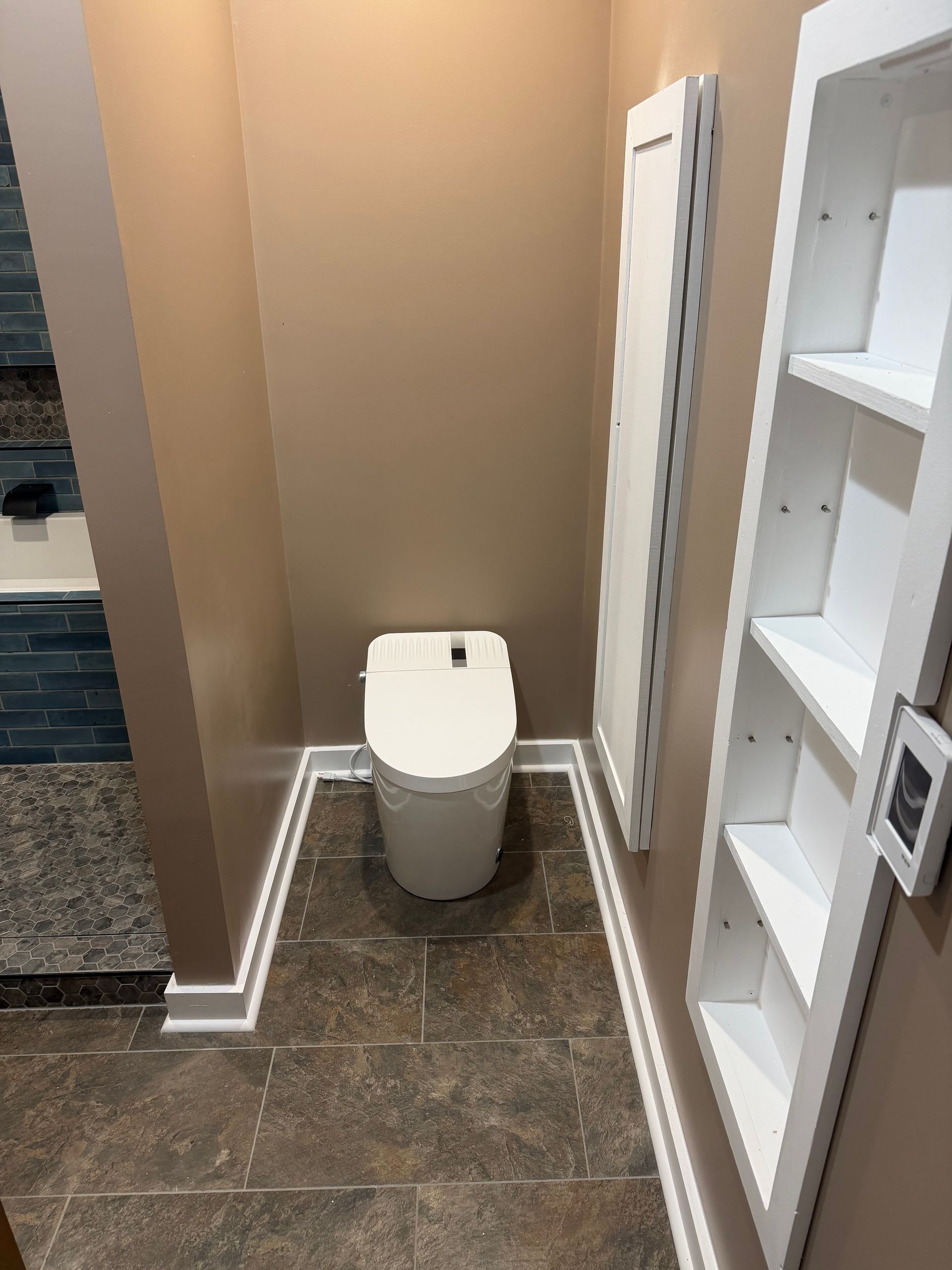 Small bathroom with a toilet centered between beige walls, dark tile floor, and white shelving on the right.