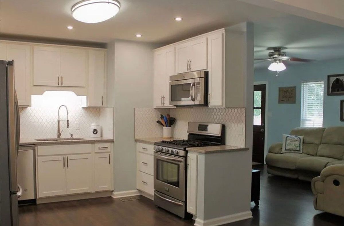 Bright kitchen with white cabinets and stainless appliances, opening to a teal living room with sofa.
