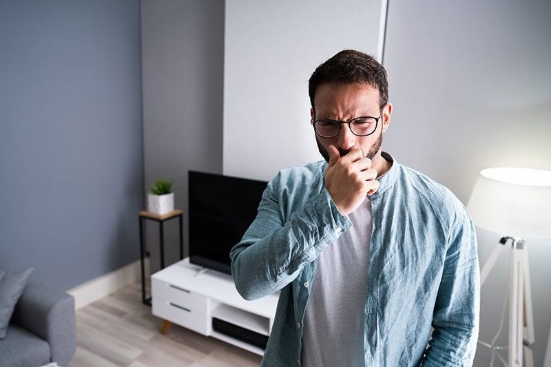 Man in a blue shirt coughing in a living room beside a TV, lamp, and couch