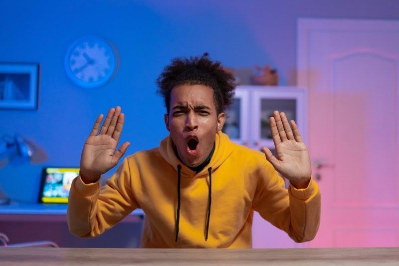 Person in a yellow hoodie shouting with hands raised at a desk in a blue-lit room.