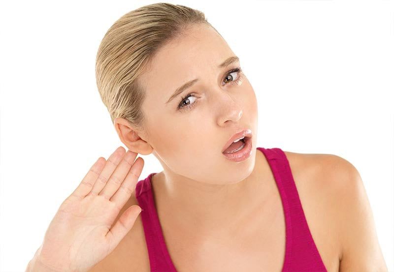 Woman in pink tank top holding a hand to her ear, looking puzzled against a white background