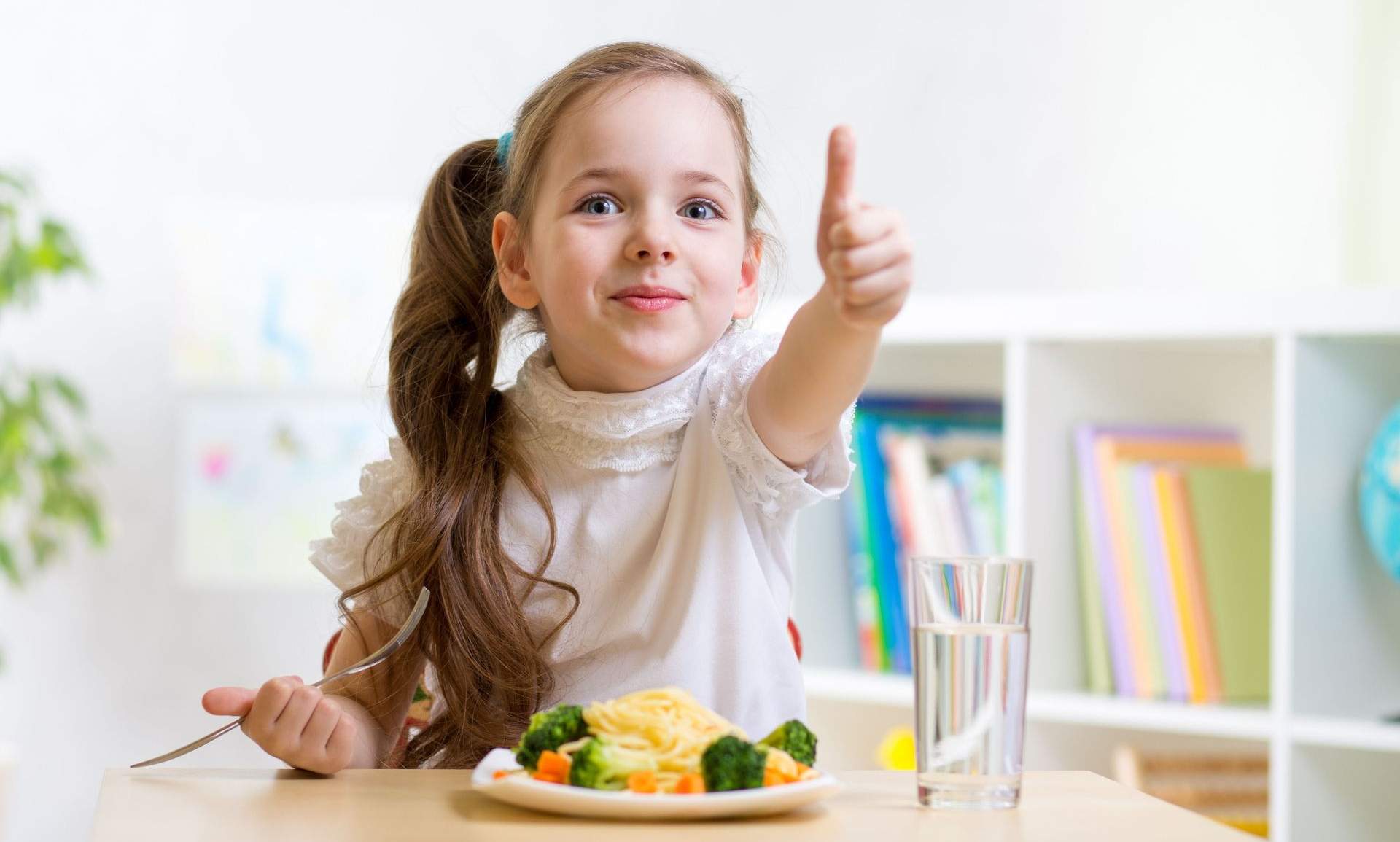 Girl giving thumbs up, with plate of vegetables and pasta, glass of water.