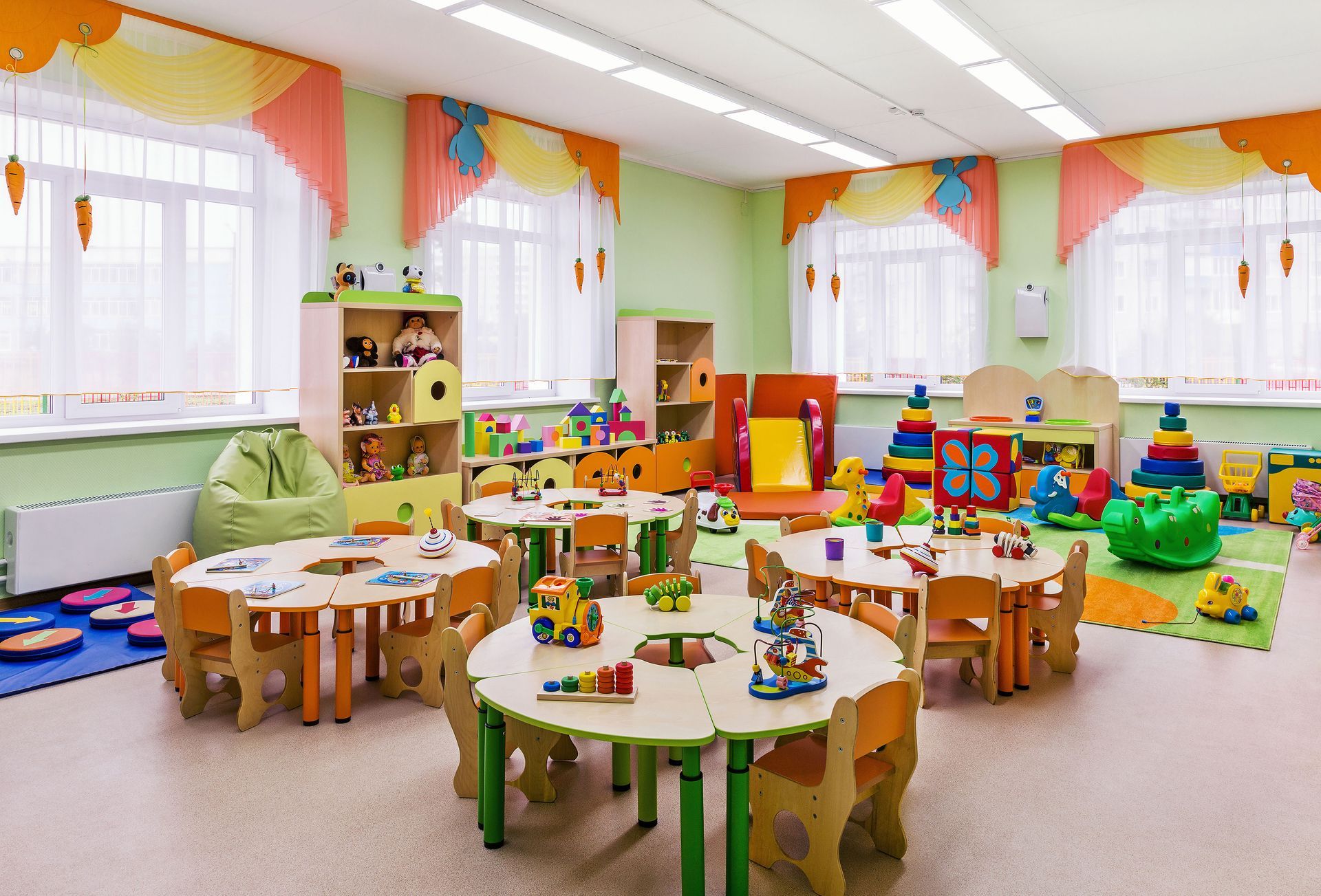 A brightly lit preschool classroom with colorful tables, toys, and shelves; windows with orange and yellow curtains.