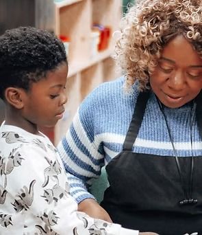 A child and adult, focused on something together. The adult wears a blue sweater and apron.