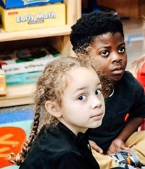 Two children looking at something off-screen. One with light hair in braids, the other with short dark hair.