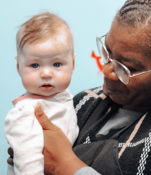 Woman holding a baby; both looking toward the camera. Baby is pale; woman is wearing glasses.