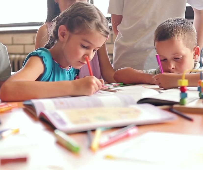 Children writing in workbooks at a table with a person standing nearby.