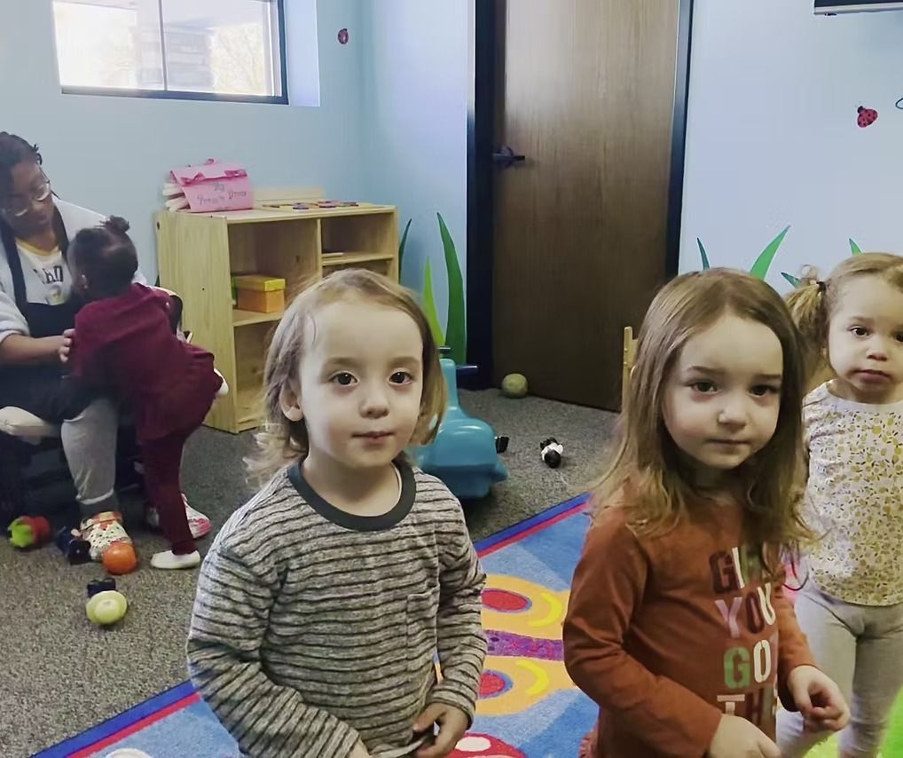 Children in a playroom looking at the camera; adult interacts with child in background.