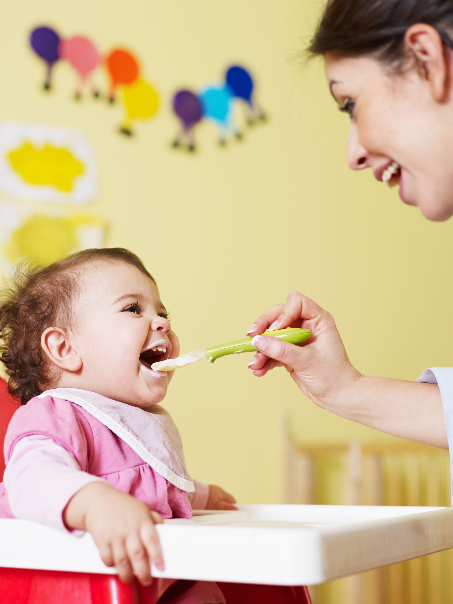 Child in high chair being fed by an adult, smiling. Playroom setting with colorful decorations.