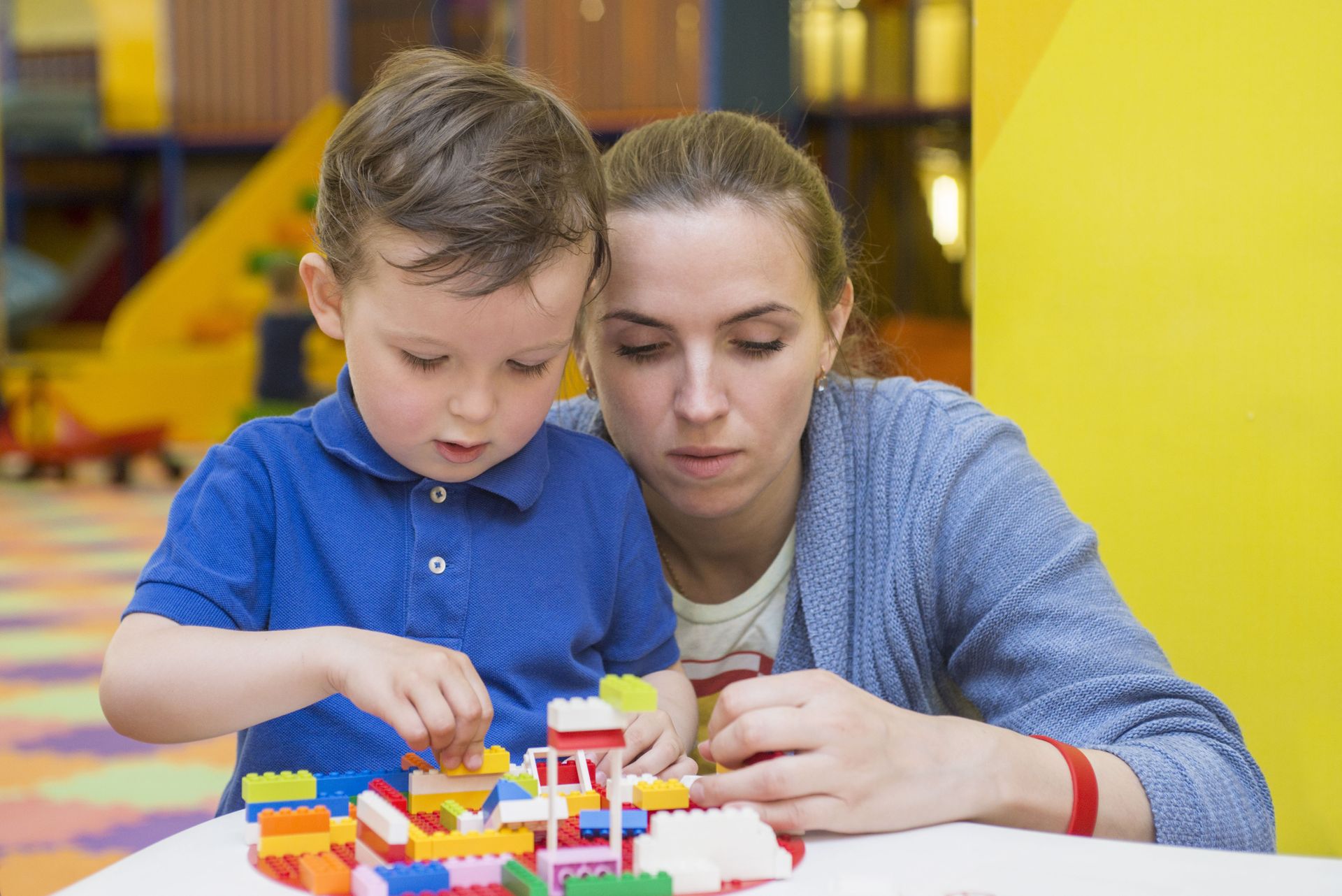 Woman and child building with colorful blocks at a table in a playroom.