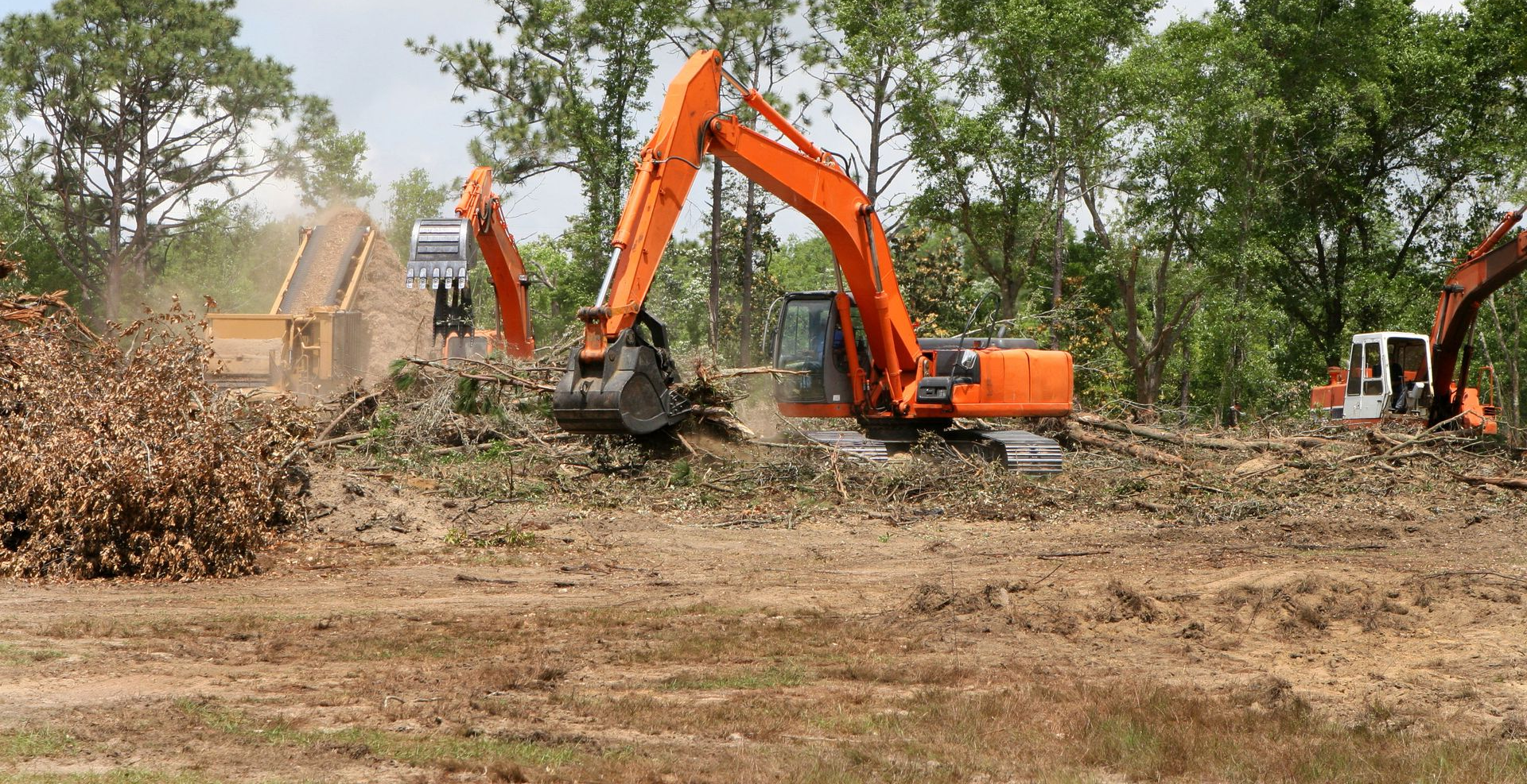 Orange excavators clearing brush in a muddy field with trees in the background