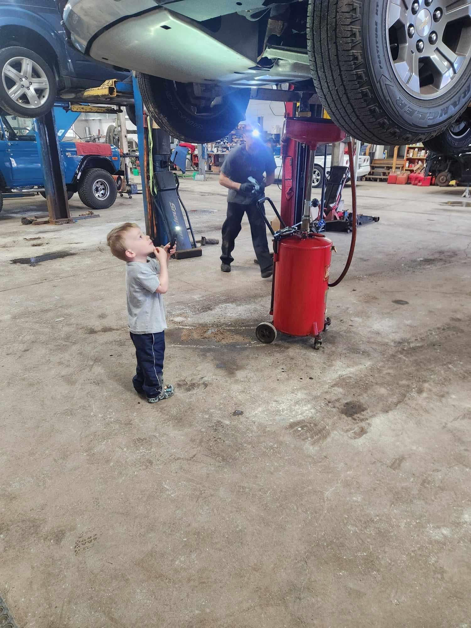 A little boy is standing in front of a car on a lift in a garage.