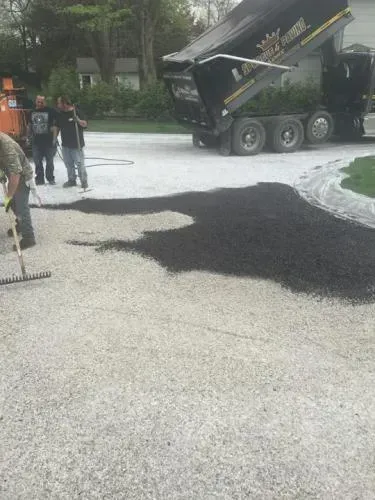 Asphalt being unloaded from a dump truck onto a driveway, with workers nearby.