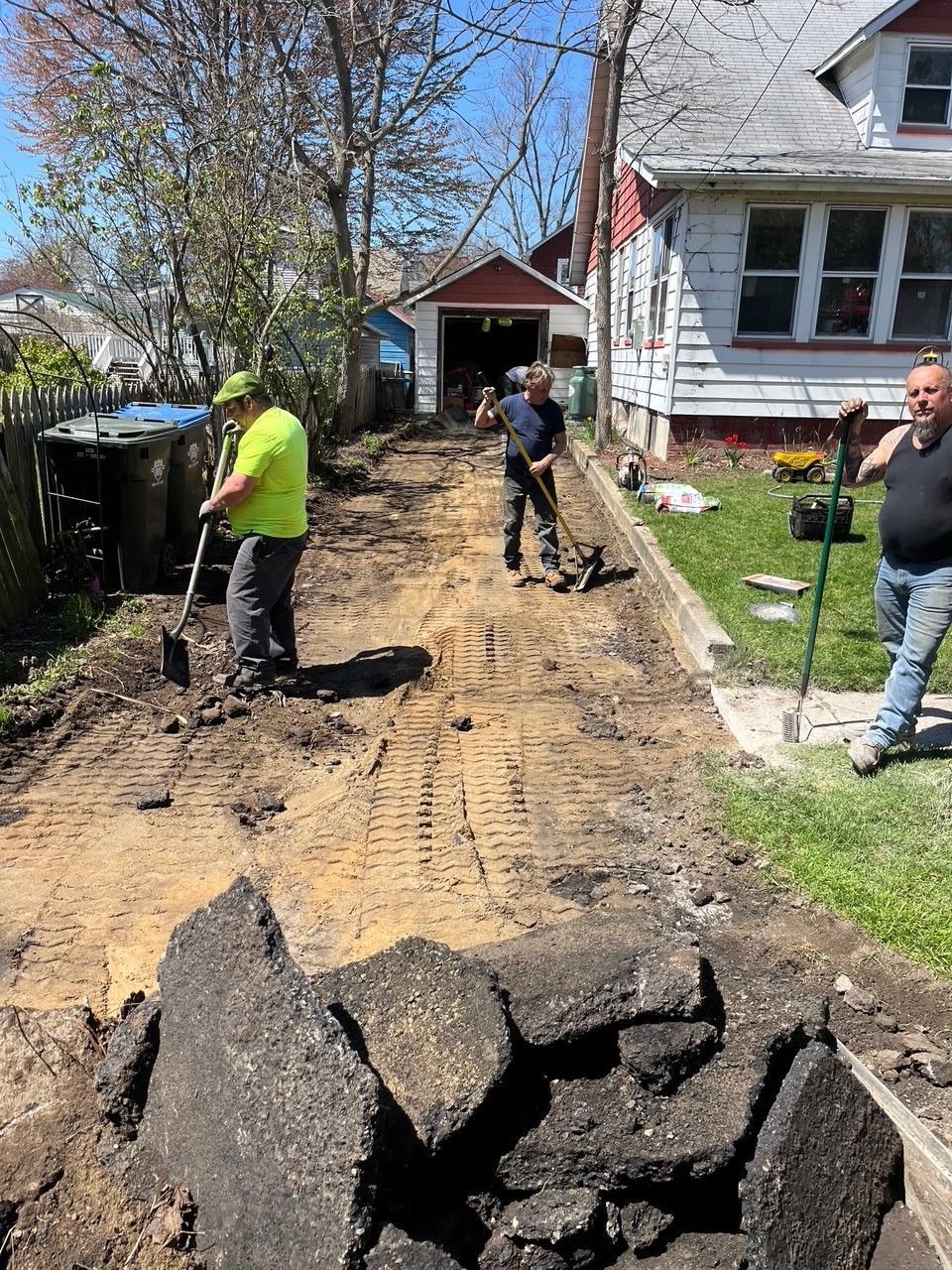 Men removing asphalt from a driveway. Piles of asphalt in the foreground. Workers use shovels, sunny day.