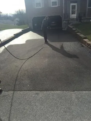 Person using a hose to clean a driveway in front of a house on a sunny day.