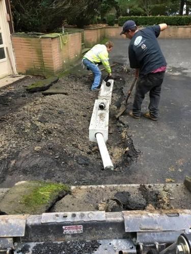Two workers installing a drainage system in a driveway. One uses a shovel, the other works with the trench.