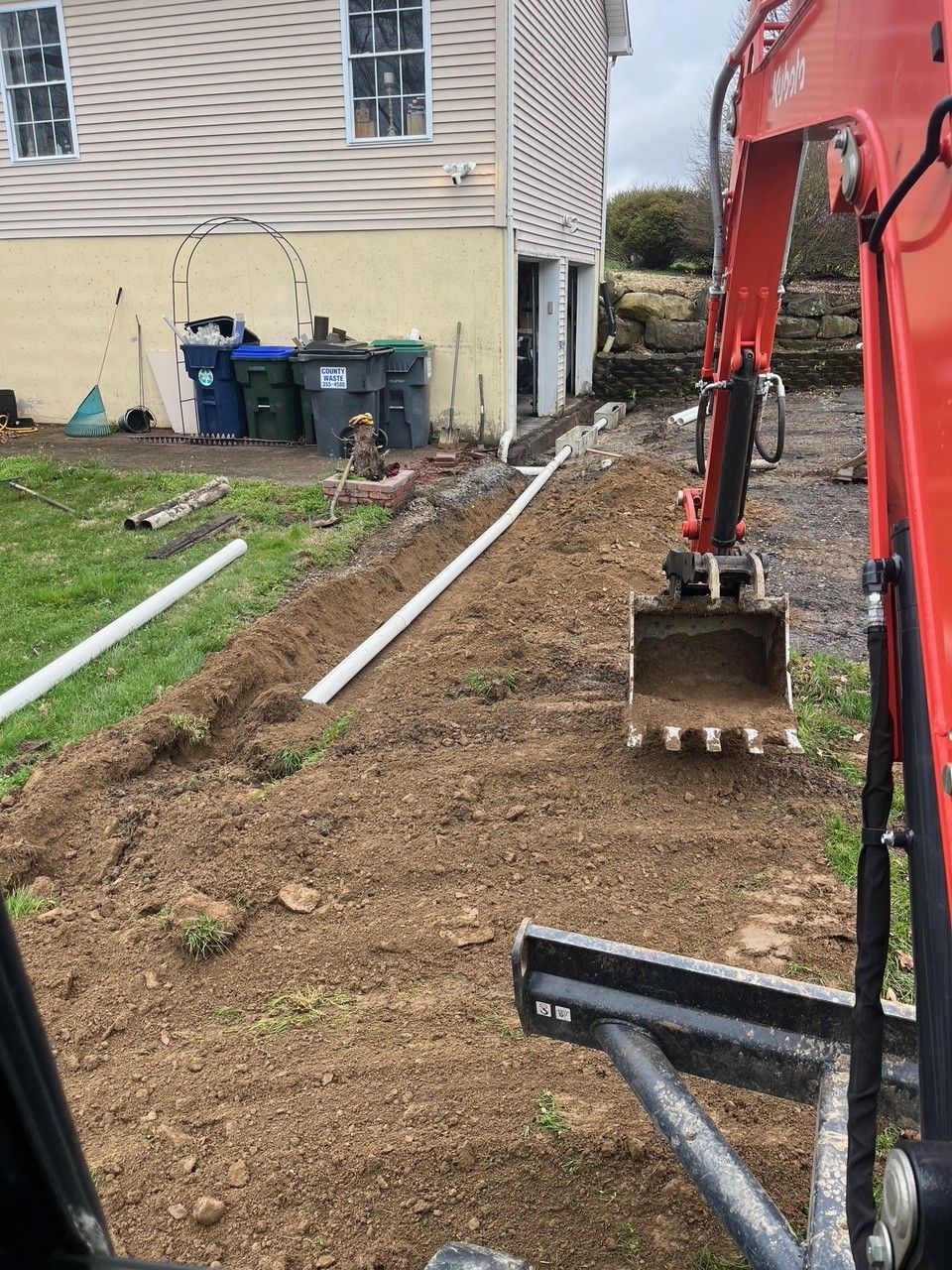 An excavator digs a trench next to a building, laying white pipes. Green grass and blue trash cans are visible.
