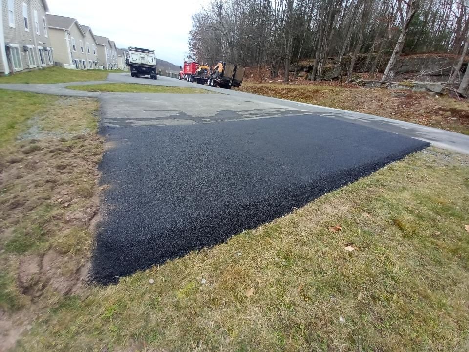 Freshly paved asphalt patch on a pathway, adjacent to grass, with construction vehicles in the background.