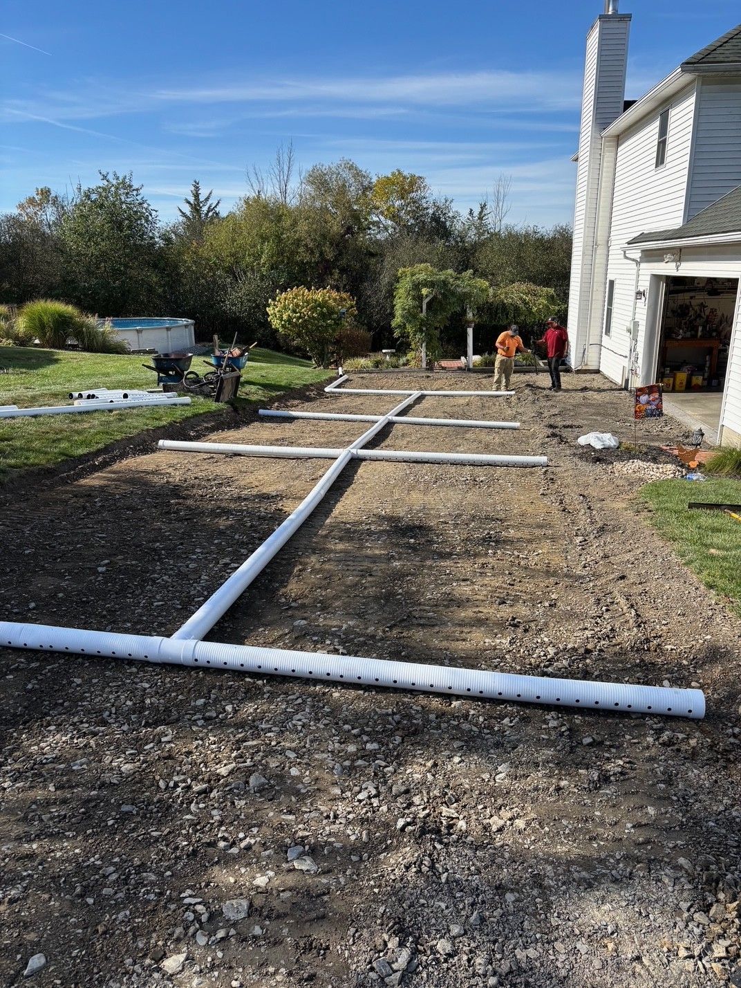 Gravel-covered yard with white pipes laid out, likely for drainage. Two workers stand near a house on a sunny day.