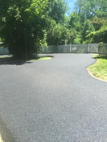 Newly paved black asphalt driveway curves towards a white fence and lush green trees.
