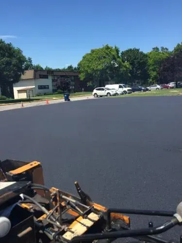 Newly paved asphalt parking lot on a sunny day with cars and a building in the background.