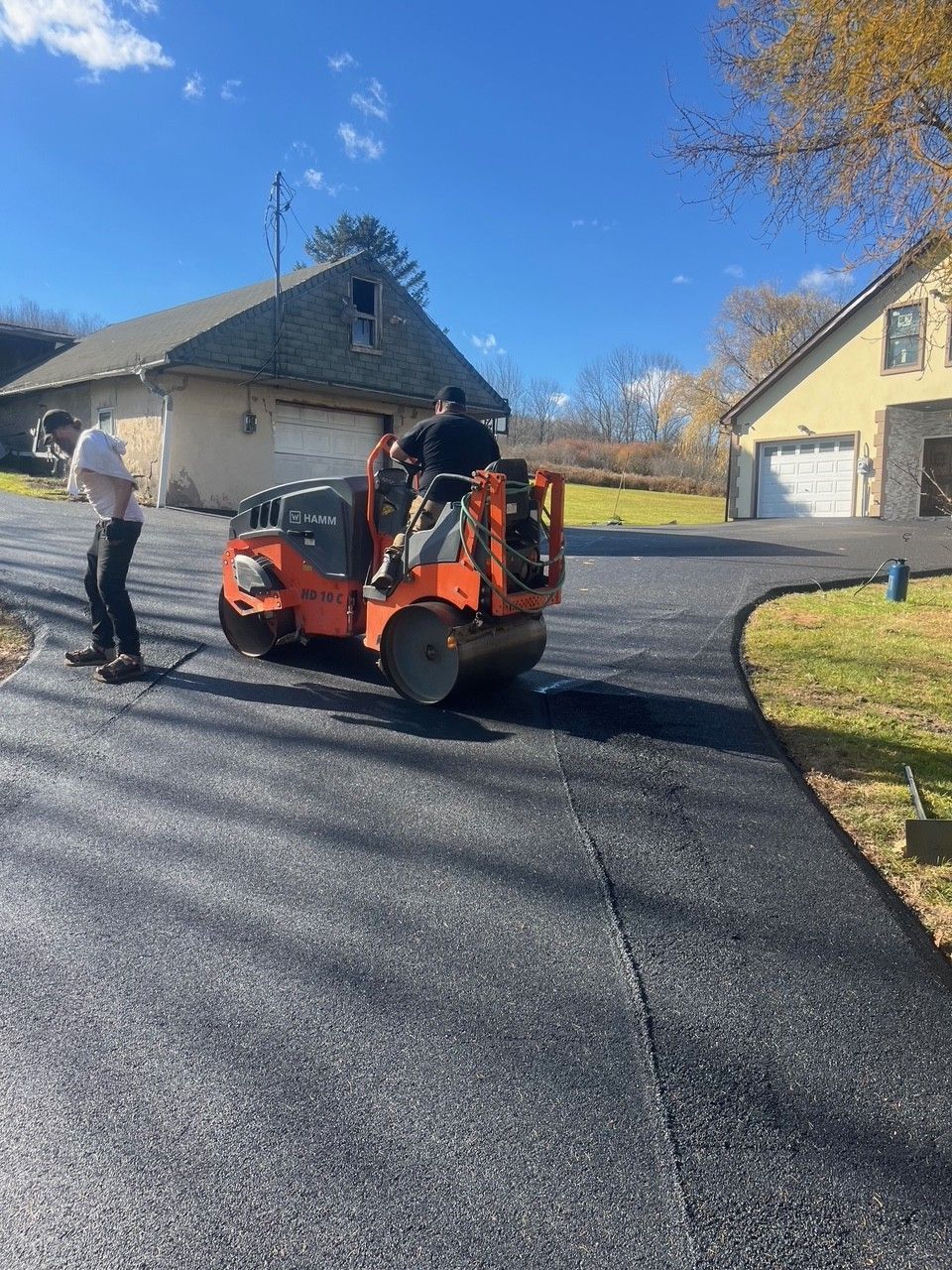 Asphalt driveway being rolled by a construction worker, with another worker nearby. Houses in background on a sunny day.