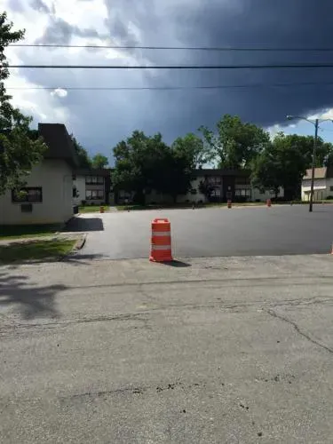 Newly paved asphalt area with an orange traffic cone, apartment buildings in the background, and a cloudy sky.