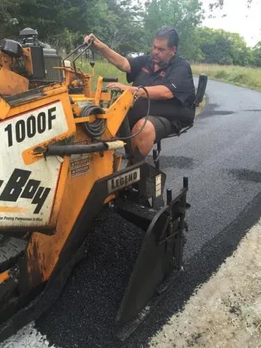 Man operating a yellow paving machine, laying fresh asphalt on a road.