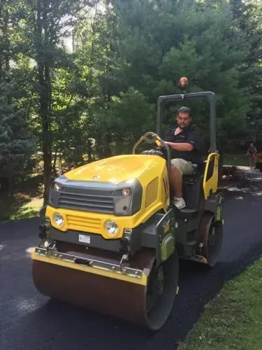 Man operating a yellow road roller on newly paved asphalt, trees in background.