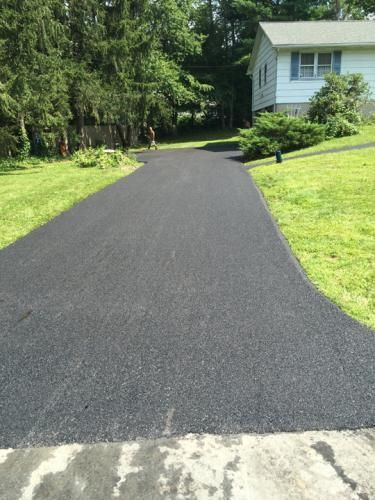 Newly paved asphalt driveway leading to a house, flanked by green grass and trees.
