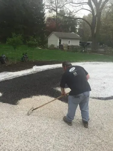 Man raking black mulch over white gravel in a yard. Small building and trees in the background.