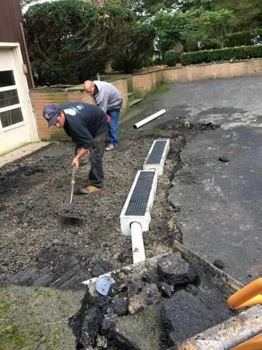 Two men install a drain in a driveway. One shovels asphalt, the other looks on. The setting is outdoors.