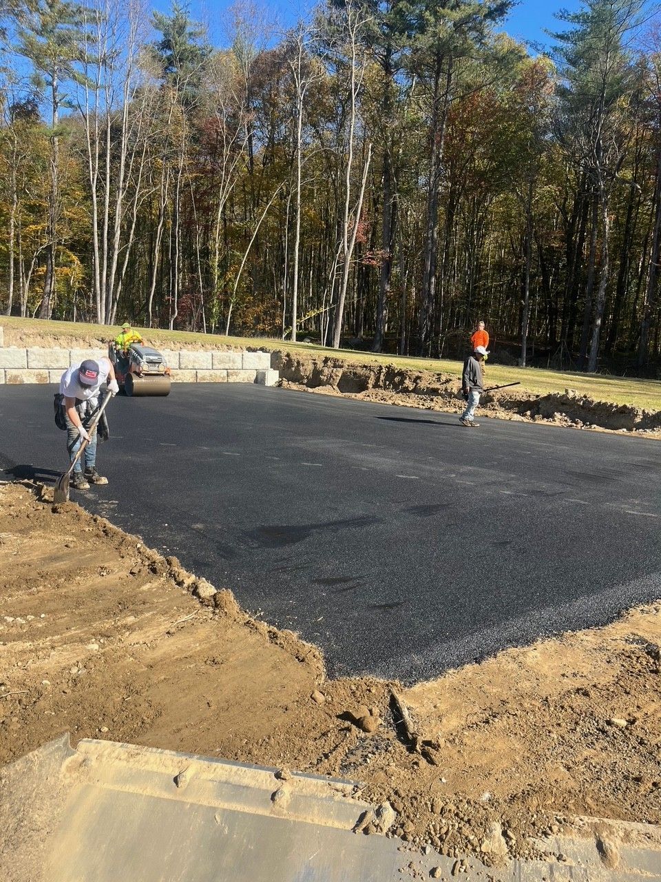 Asphalt paving in progress: workers, roller, fresh blacktop, dirt edges, trees in background.
