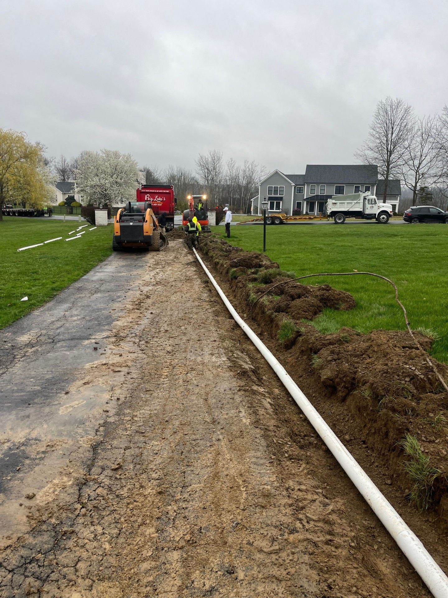 Construction crew installing a white pipe in a trench along a driveway. Heavy machinery and workers are present.