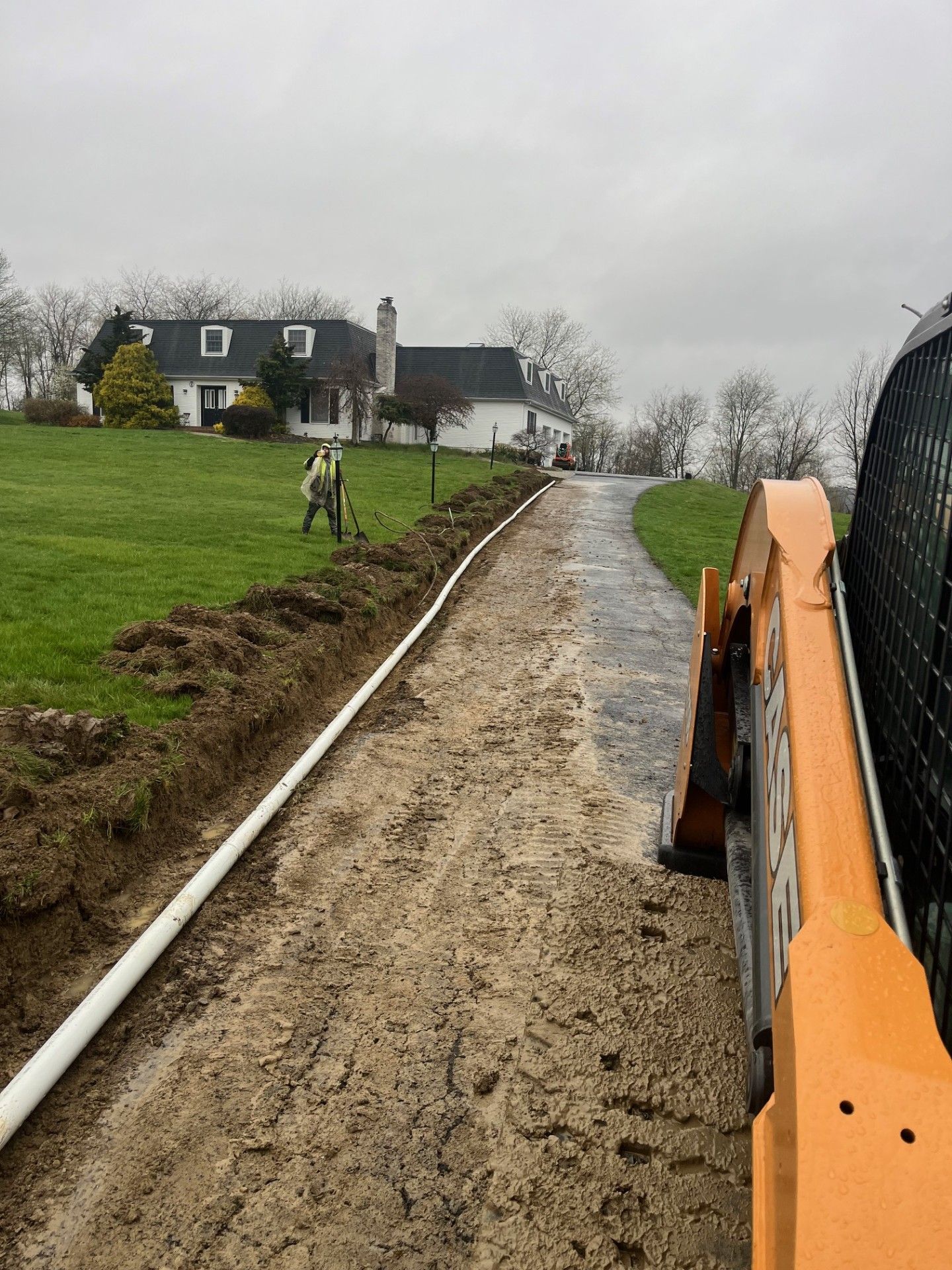 Construction site: Trench with white pipe, asphalt driveway, person digging, and house.