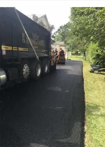 Asphalt paving in progress: truck unloading, roller compacting, newly laid black surface alongside grass.