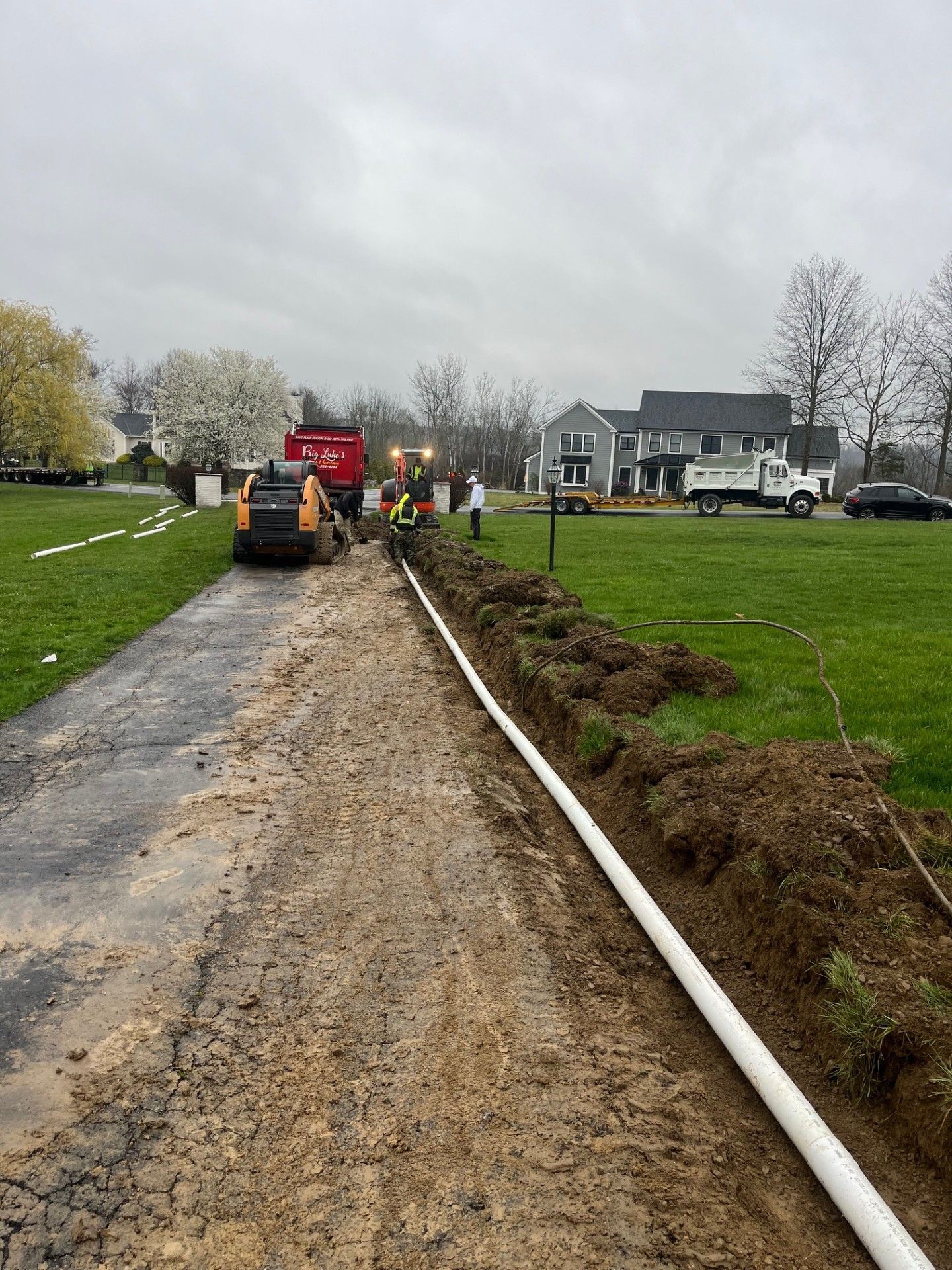 Construction crew working on a trench along a driveway, white pipe installed, overcast sky.