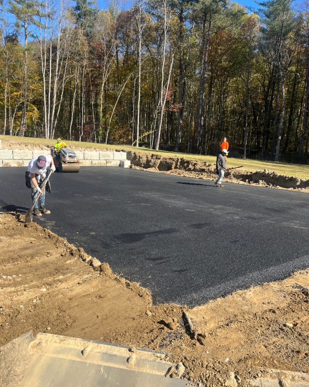 Workers paving a black asphalt surface with a roller and shovels, dirt edges in a wooded area.
