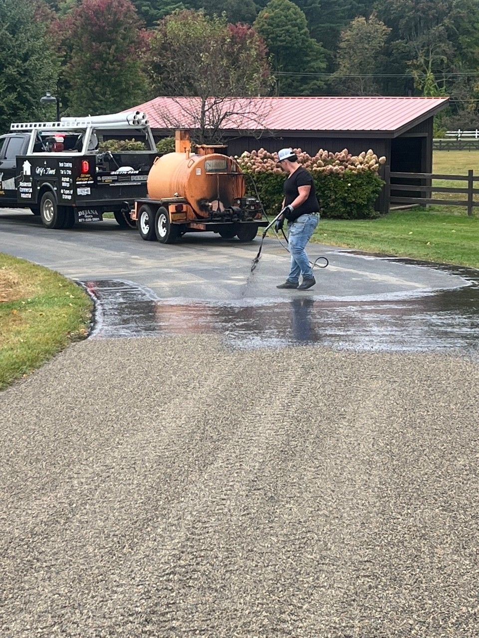 Person sealcoating a driveway with a trailer-mounted tank and a spray wand.