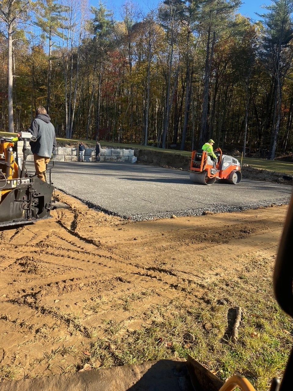 Construction workers paving a gravel lot with machinery. Autumn trees in the background.