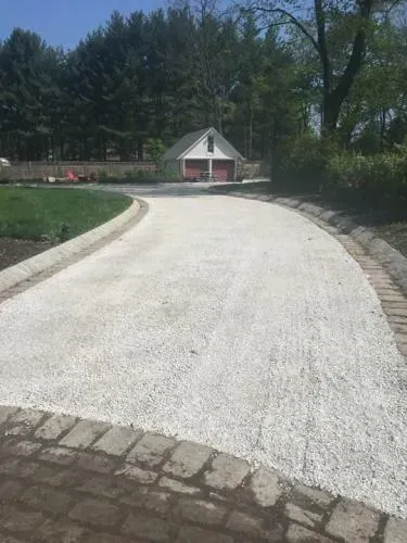 A white gravel driveway curves towards a small red and white building, bordered by brick and greenery.