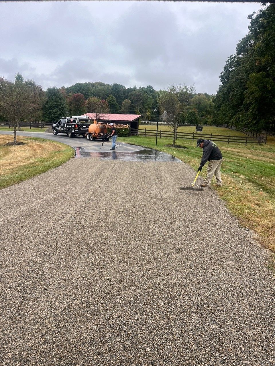 Man rakes gravel driveway, a truck and workers in the background. Cloudy day.