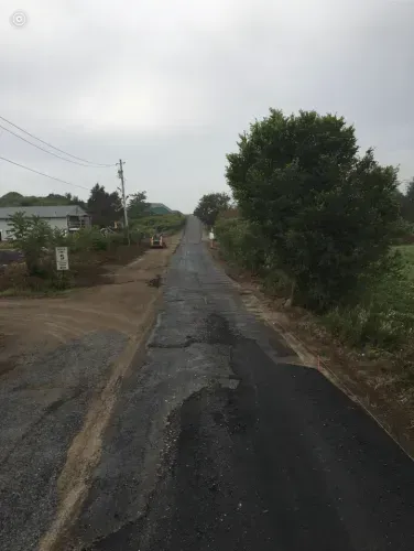 Paved road with repaired sections, leading uphill toward cloudy sky, houses on the left, trees on the right.
