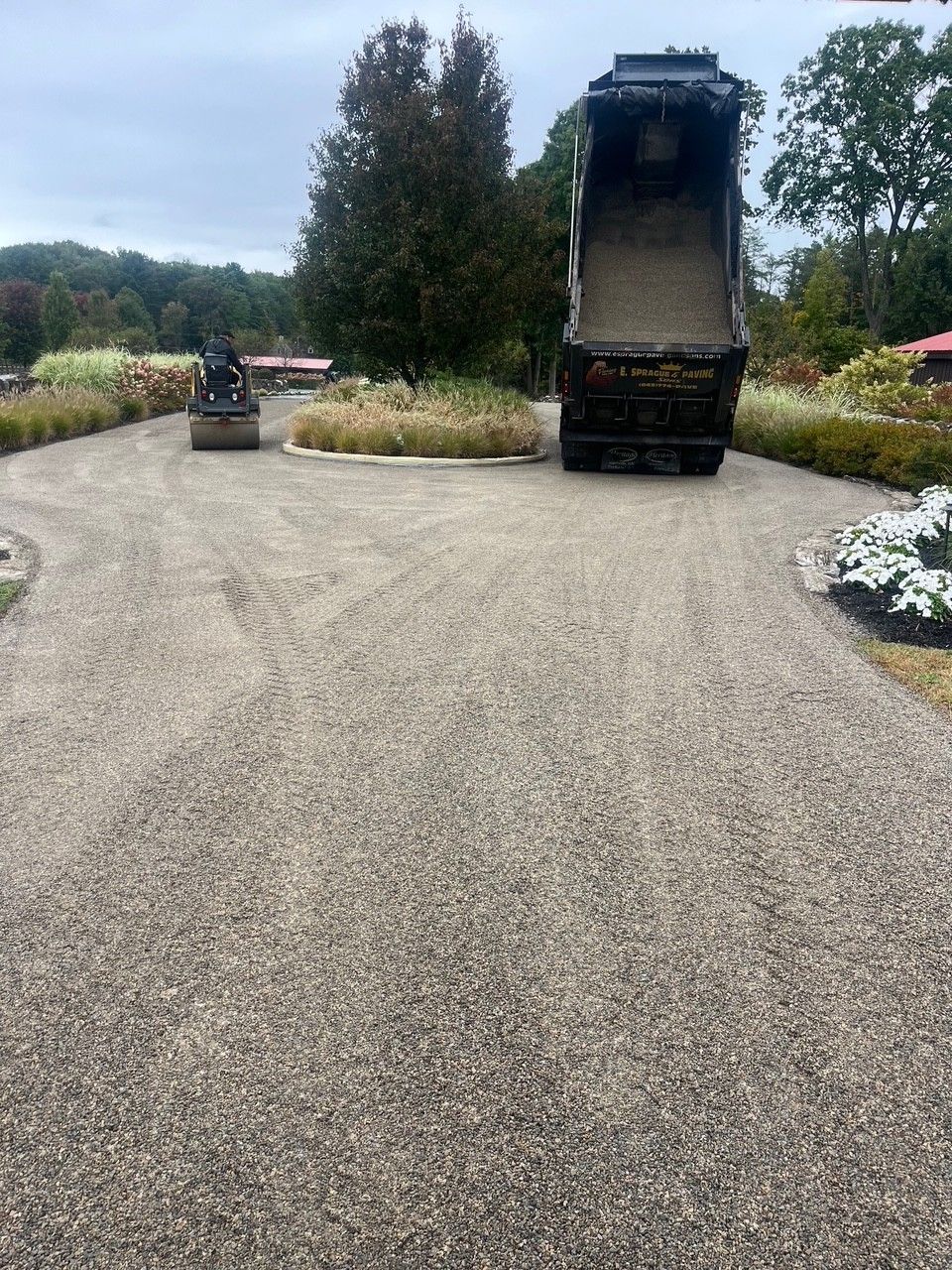 A dump truck unloading gravel onto a garden path next to a landscape with a roller compactor in the distance.